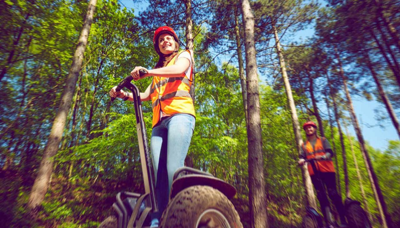 Two riders operate off-road Segways, leaning on handlebars while moving along a dirt trail. They wear red helmets and orange safety vests. Green trees surround them under a blue sky.