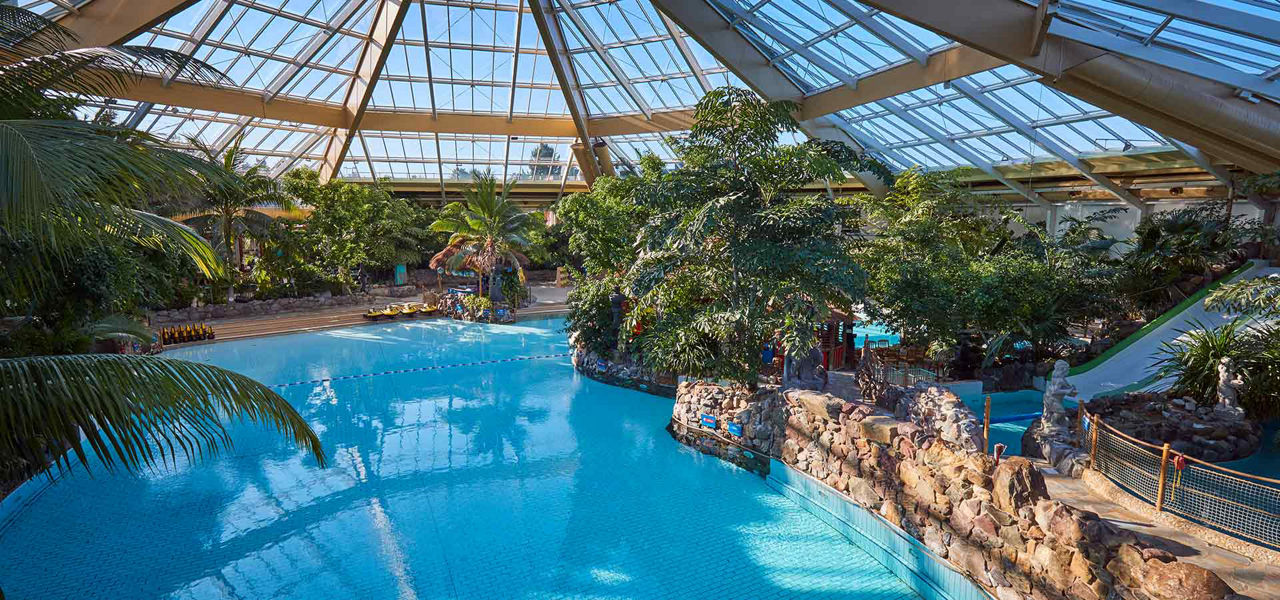 Large indoor swimming pool reflects sunlight, surrounded by tropical plants and rock-lined channels; waterslide curves down right side under a glass-domed roof, with lounge chairs and pathways around the water.