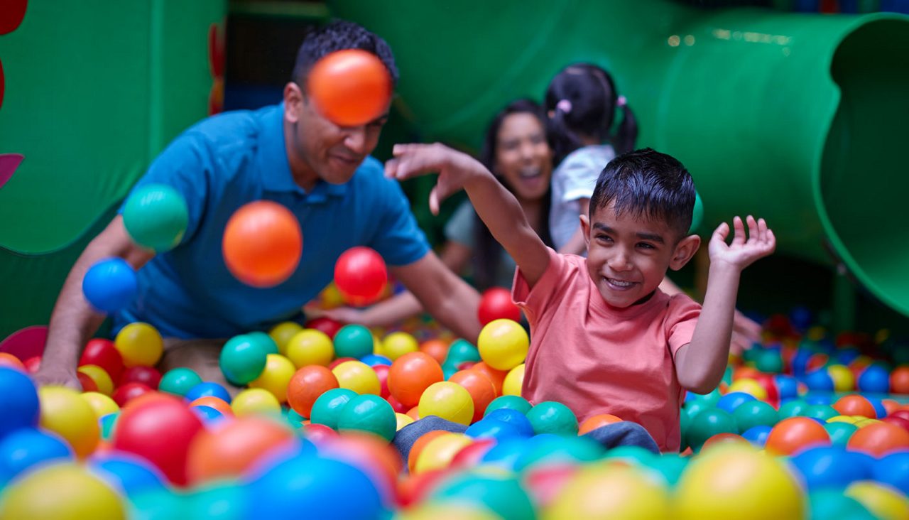 Child smiles and tosses colorful plastic balls, surrounded by bouncing balls; nearby adults play and watch in background; indoor play area with a ball pit and a green tube slide.
