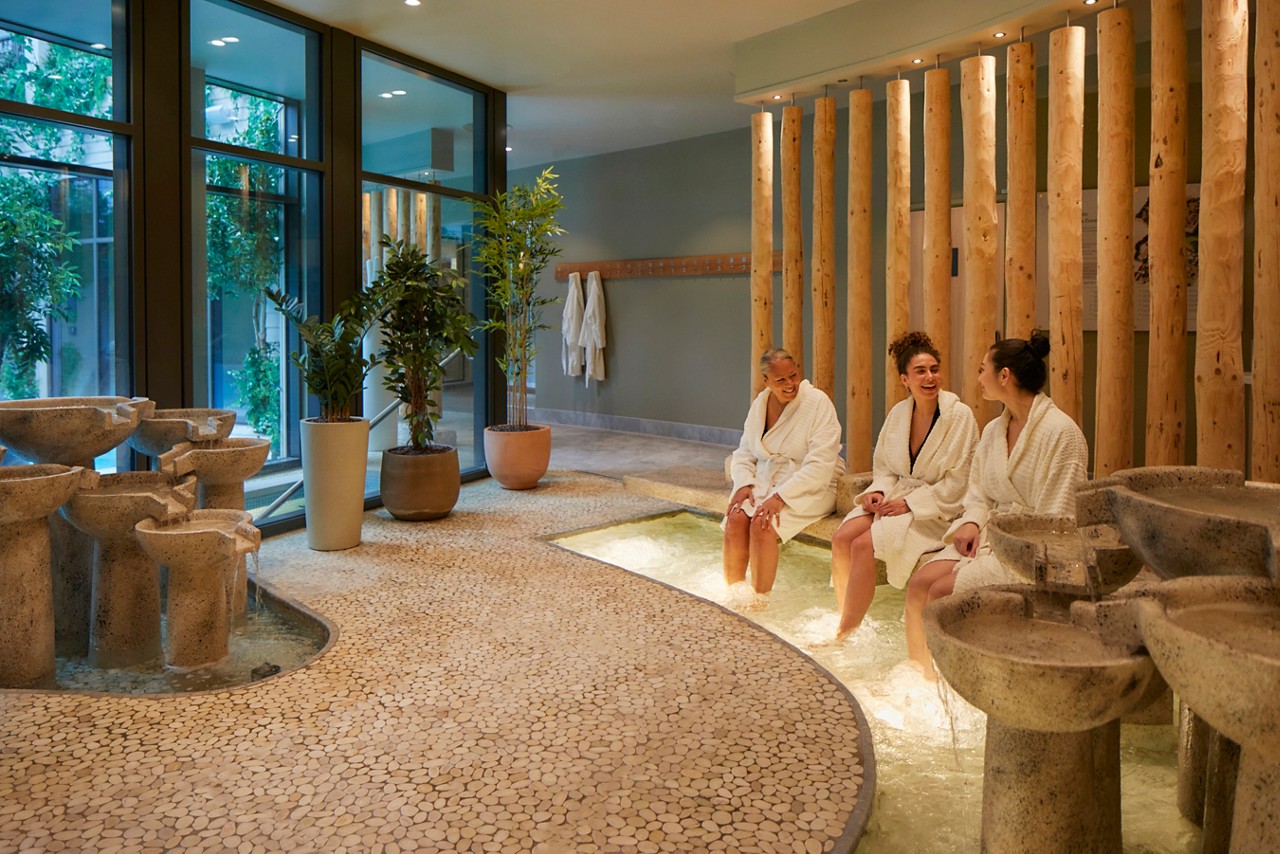 3 women sat on a bench in spa robes with their feet in a foot bath