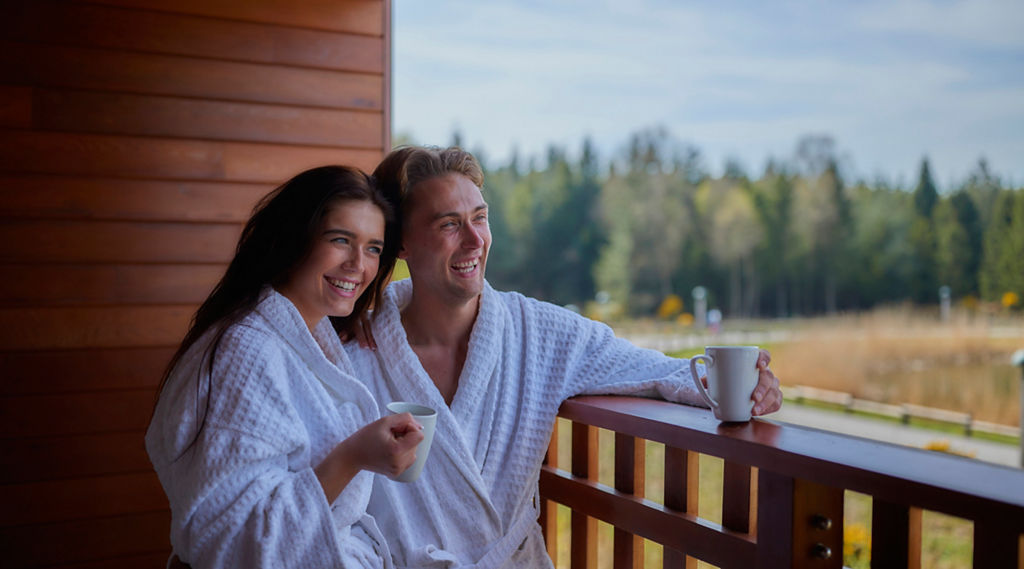 Two people in white bathrobes smile and hold mugs, leaning on a wooden balcony railing, overlooking a lakeside meadow and distant trees under a bright, partly cloudy sky.