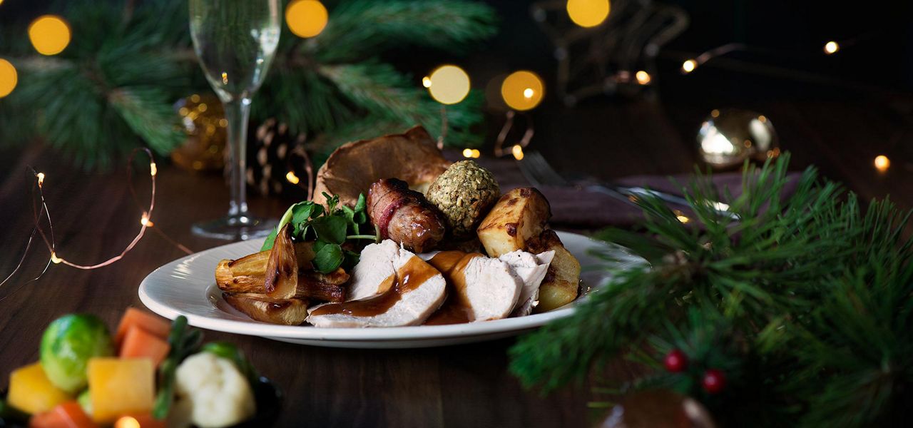 Roast turkey slices with gravy sit on a plate beside roast potatoes, stuffing, bacon-wrapped sausage, and greens, set on a festive table with pine, fairy lights, and a champagne flute.