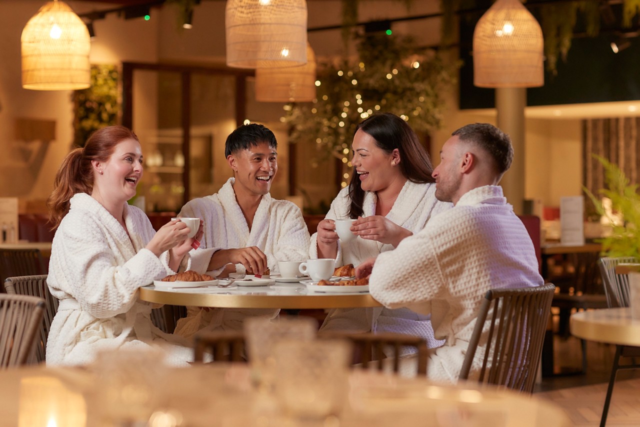 a group of friends sat at a table in spa robes laughing and enjoying food