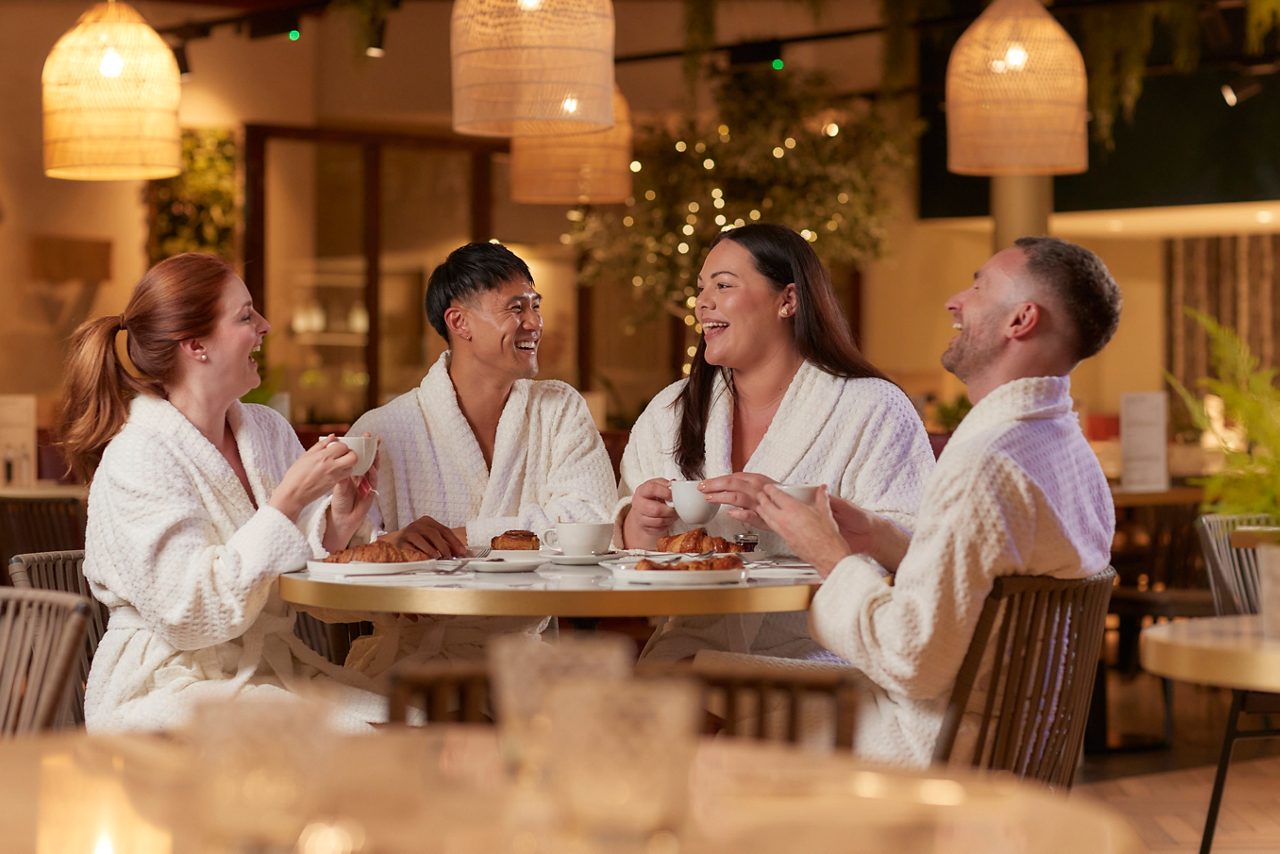 four people in spa robes sat at a table laughing while enjoying food and drink