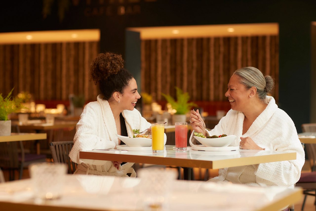 Two people in white robes eat and chat over salad bowls and orange and pink juices, seated at a modern indoor cafe with warm lighting, plants, and wooden accents.