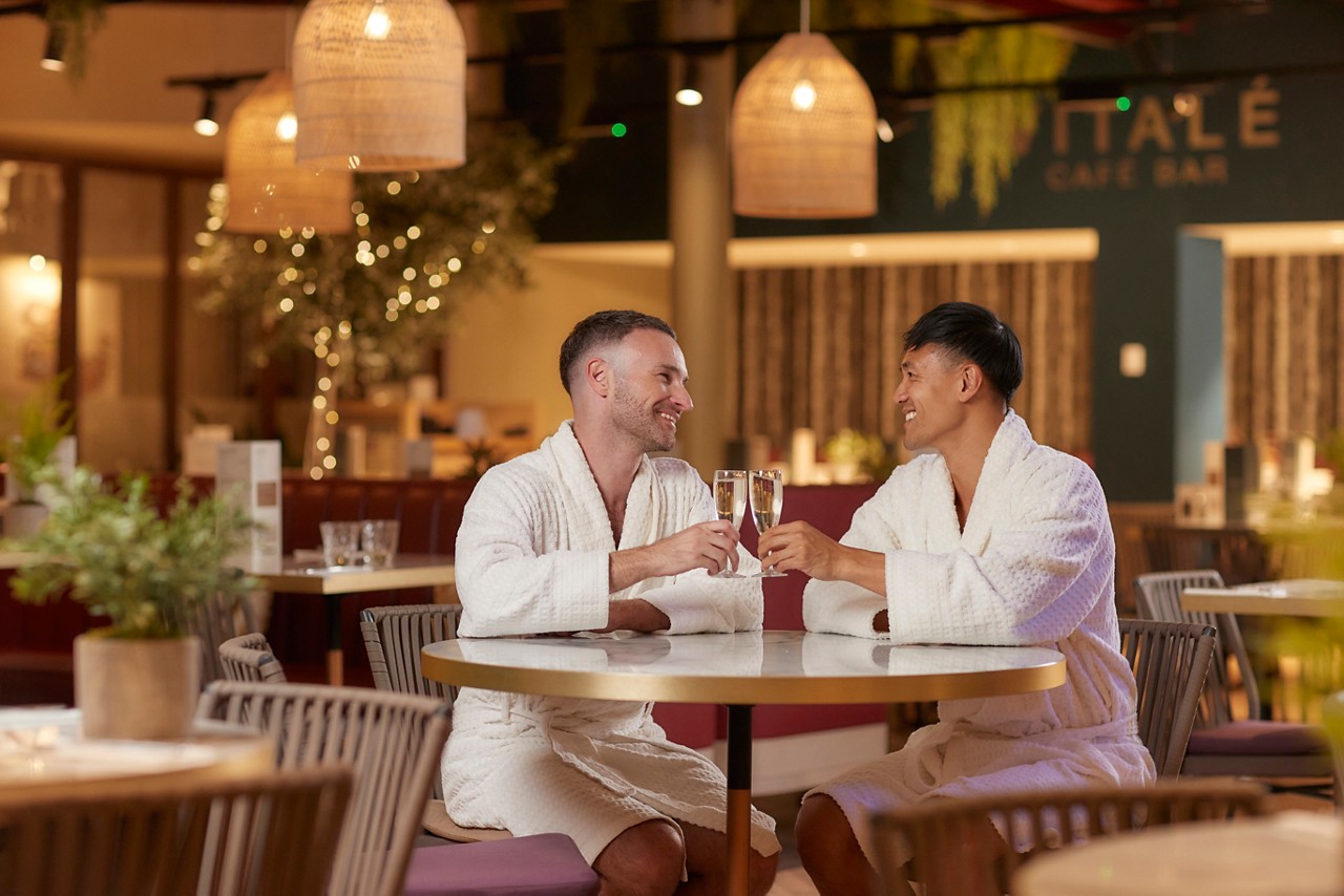 two people in spa robes sat at a table with glasses of prosecco