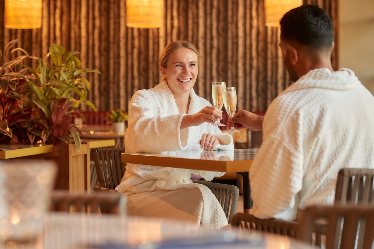 A couple is spa robes sat at a table clinking glasses of prosecco