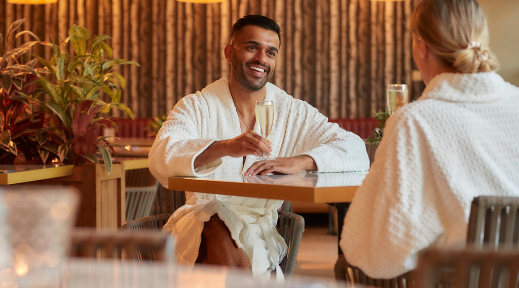 a man and women sat at a table in spa robes with glasses of prosecco