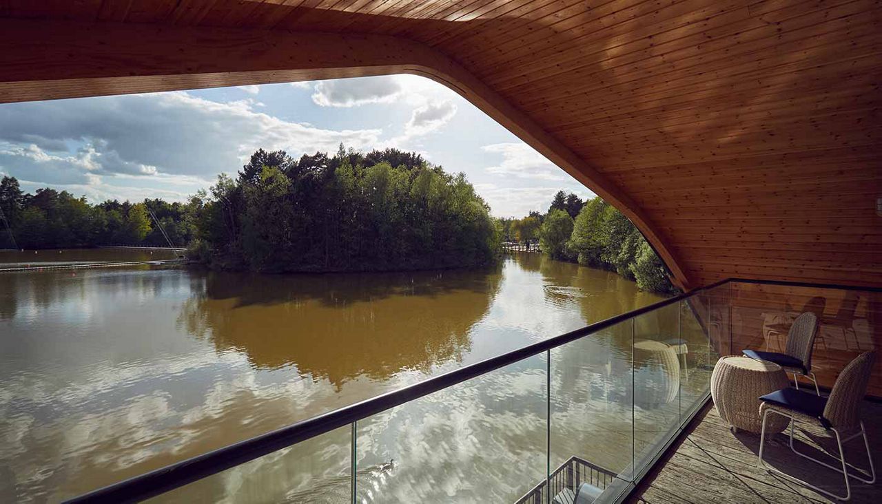 Balcony with glass railing overlooks a calm, reflective lake; two chairs and a small table sit beneath an arched wooden roof, with tree-lined shores and a partly cloudy sky beyond.