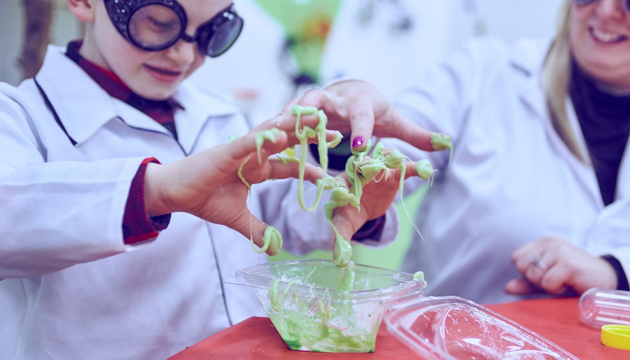young boy in scientist jacket and goggles sticking fingers into green slime