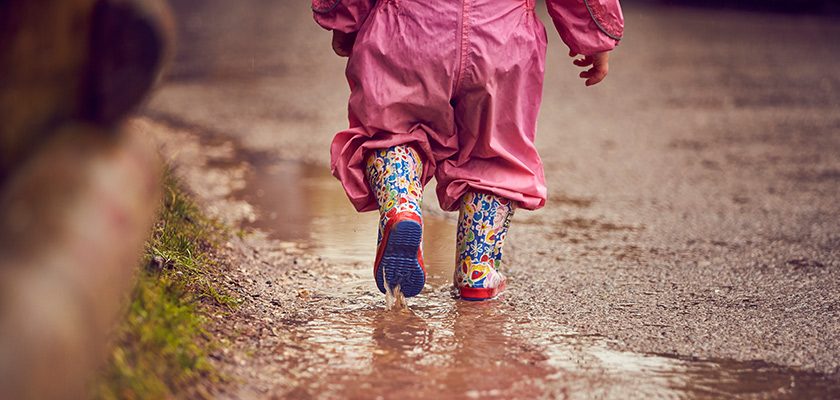Child in a pink rain suit steps through a shallow puddle, colorful rubber boots splashing water, on a wet path bordered by grass and wooden posts.