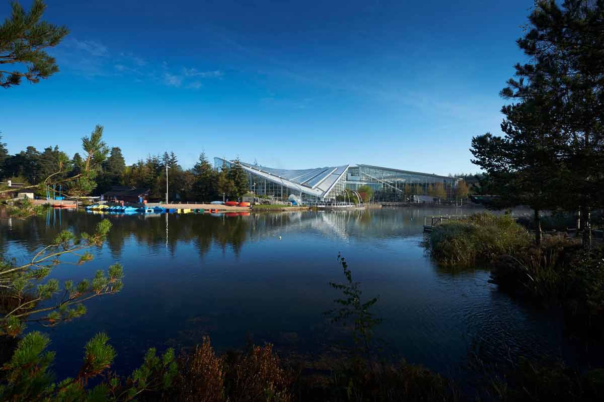 Glass-roofed leisure complex reflects on a calm lake, flanked by forest. Colorful paddle boats line a dock; pine trees and reeds frame the scene beneath a clear blue sky.