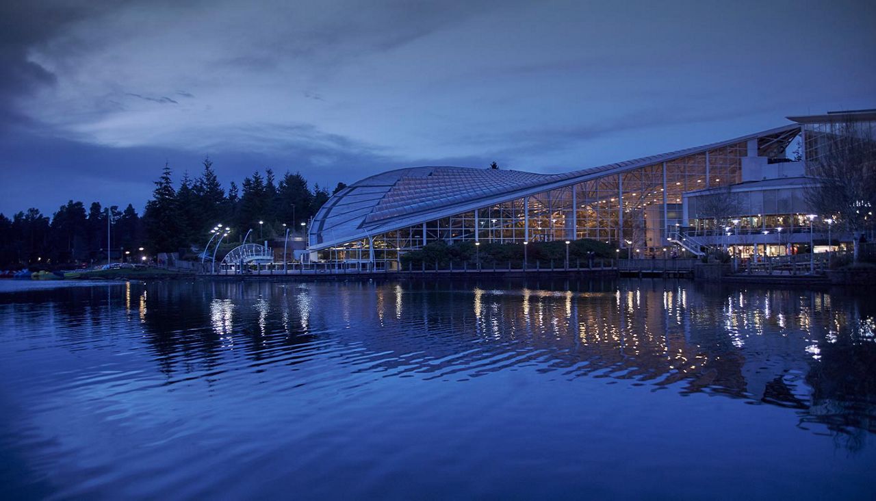 A large, curved glass-roof building glows warmly, casting reflections across calm lake water; pedestrians and lampposts line a waterfront boardwalk beside trees under a deep blue evening sky.