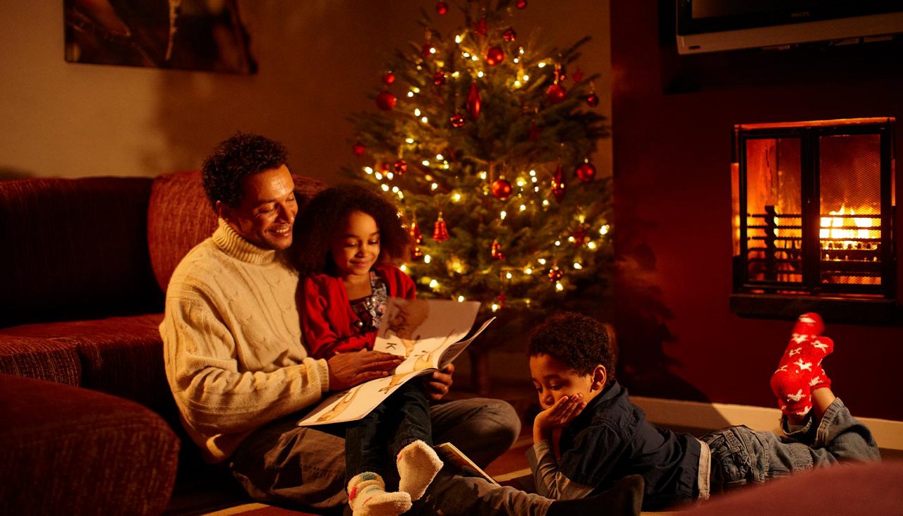 family sat by the christmas tree reading a story book together. The fire is glowing in the back and the room is softly lit by christmas tree lights